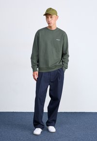 Sudadera verde, pantalones de rayas oscuros, zapatillas blancas y una gorra verde. Look informal con tejido texturizado y logo bordado en la sudadera.