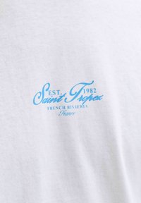 Biela tkanina s modrým textom „EST. 1982 Saint Tropez French Riviera France“ písaným kurzívou a veľkými písmenami.