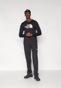 Μαύρο πουκάμισο με μακριά μανίκια και παντελόνι, και τα δύο με το λογότυπο "The North Face" σε άσπρο. Μαύρο καπέλο και μαύρα παπούτσια ολοκληρώνουν το σύνολο.