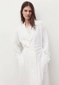 Rochie albă cu mâneci lungi, cu guler tip șal, cu front butonat și talie legată. Fabricată dintr-un material moale cu o textură netedă și cu buzunare.