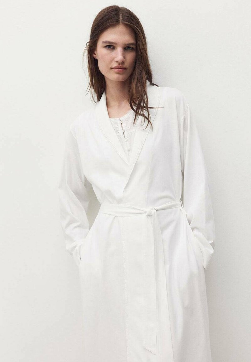 Rochie albă cu mâneci lungi, cu guler tip șal, cu front butonat și talie legată. Fabricată dintr-un material moale cu o textură netedă și cu buzunare.