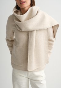 Sieviete valkā bēša vilnas jaku ar lielu atbilstošu šalli, kas pārmetta pāri vienam plecam, un baltas bikses uz vienkāršas fona.