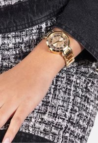 Zlaté analógové náramkové hodinky s okrúhlym puzdrom, krystalovými akcentmi na ciferníku a zlatým článkovým náramkom, nosené na svetlej pleti zápästí.