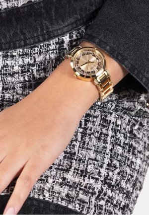 Zlata analogna uro z okroglo ohišjem, kristalnimi akcenti na številčnici in zlatim zapestnim naramkom, nosi jo oseba s svetlo kožo.