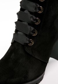 Botte de cheville en suède noir avec des lacets en satin passés à travers des œillets en or, présentant un talon épais et une surface texturée.