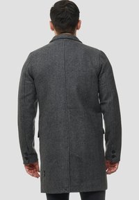 Grå uldblandet frakke med tekstureret overflade, med en hakket krave, sidelommer og manchetter med knapper. Baksiden viser et strømlinet design.