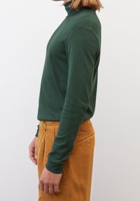 Maglione a collo alto verde a maniche lunghe in un morbido tessuto, con una forma aderente, indossato con pantaloni in corduroy arancioni. Mostra una trama liscia e un design semplice.
