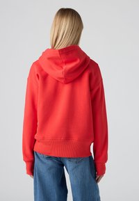 Persoană cu păr lung blond, purtând un hanorac roșu aprins și blugi, întoarsă cu fața la un fundal deschis.