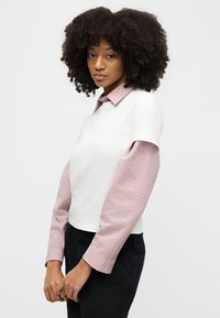 Γυναίκα με σγουρά μαλλιά που φοράει λευκή κοντομάνικη μπλούζα πάνω από ροζ μπλούζα μακρυμάνικη με υφή, όρθια με τα χέρια της ενωμένα.