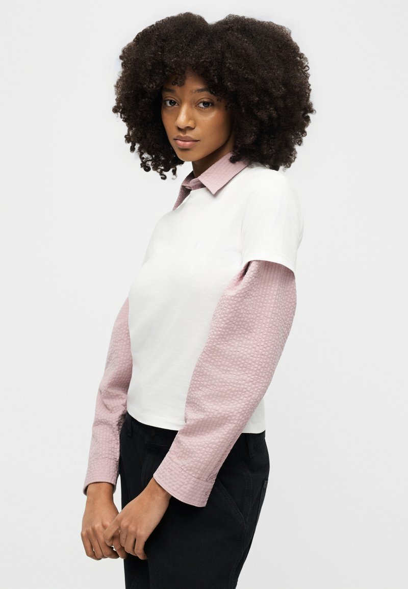 Donna con capelli ricci che indossa una maglietta bianca a maniche corte sovrapposta a una camicia rosa a maniche lunghe e strutturata, in piedi con le mani intrecciate.