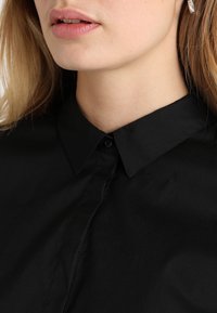 Chemise noire à col avec une texture lisse, dotée d'une fermeture à boutons et d'un design classique. Le tissu semble léger et formel.