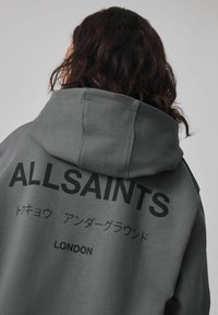 Felpa con cappuccio grigia con grande testo nero "ALLSAINTS" e altre lingue sul retro, caratterizzata da una texture di tessuto morbido e un cappuccio con cordino.