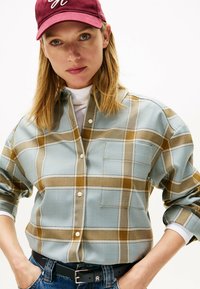Chemise à carreaux bleue et marron avec une poche sur la poitrine, design à boutons, et manches retroussées, portée avec un col roulé blanc et une casquette bordeaux.