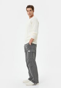 Suéter de punto crema claro y pantalones cargo grises con bolsillos laterales, pasadores para cinturón y una etiqueta. El modelo está de pie con las manos en los bolsillos, usando zapatos blancos.
