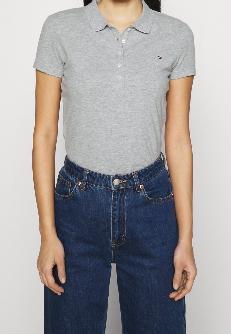 Personne portant un polo cintré à manches courtes gris clair avec patte de boutonnage et un jean taille haute bleu foncé, debout les bras détendus.
