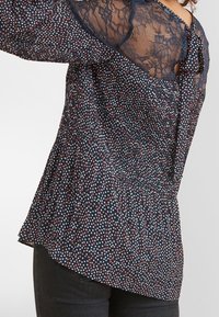 Blusa blu navy con pois multicolori, schiena in pizzo, maniche lunghe a sbuffo e vita arricciata. Tessuto morbido e testurizzato, con chiusura a bottoni.
