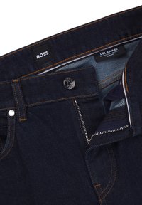 Dark blue denim jeans featuring a slim fit, signature logo on the waistband, contrasting orange stitching, and metallic zipper closure.