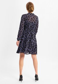 Schwarzes Blumenkleid mit langen Ärmeln, das ein fließendes Design und kleine lila Blumen aufweist. Getragen mit schwarzen High Heels.