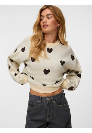 Donna con capelli biondi lunghi che indossa un maglione crema con motivi a cuore neri e jeans scuri a vita alta, posando con le mani sui fianchi.