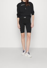 Svart kortad hoodie med logotyp på bröstet, kombinerat med svarta cykelshorts med logotypdetalj. Modellen bär vita sneakers med svarta accenter.