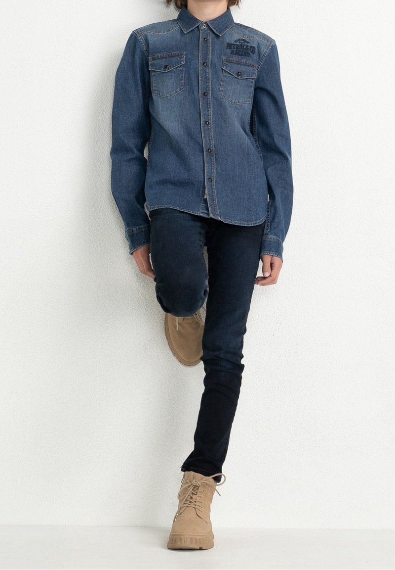 Chemise en denim bleu moyen avec un col, deux poches poitrine et fermeture à boutons, associée à un jean noir et des bottes montantes beiges.