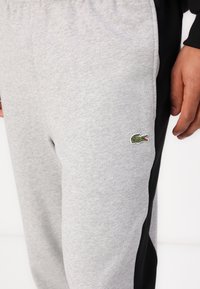 Pantaloni de trening gri din bumbac cu panouri laterale negre. Prezintă un logo brodat cu un crocodil verde aproape de șoldul stâng. Textură moale, talie elastică.