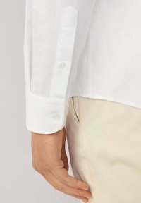 La mano sinistra dell'uomo è rilassata accanto al corpo, indossa una camicia bianca a maniche lunghe con polsini bottonati e pantaloni beige con tasca laterale visibile.