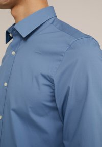Modra srajca na gumbe s ostrim ovratnikom, izdelana iz gladkega materiala. Ima subtilen sijaj in standardno zapiranje z gumbi.
