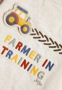 Vaaleanharmaa puuvillapaita, jossa on värikäs "FARMER IN TRAINING" -brodeeraus ja traktori-logo; sisältää ruskeita koristeompeleita.
