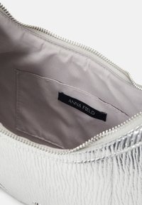 Srebrna metaliczna saszetka o teksturowanej powierzchni, zapinana na zamek błyskawiczny, z gładkim beżowym wnętrzem. Ozdobiona etykietą z napisem "ANNA FIELD".