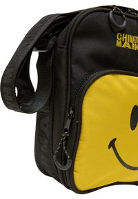 Sort stof crossbody tas med et gult smiley ansigt design, tekstureret frontlomme, justerbar rem og sorte lynlåsdetaljer.