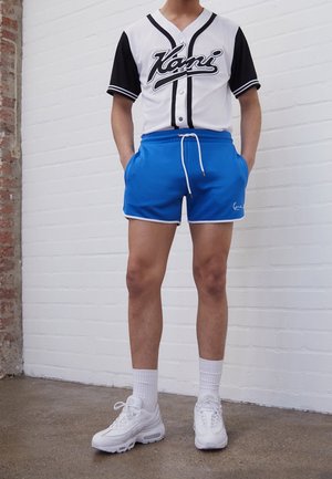 Weißes und schwarzes Baseball-Trikot mit kurzen Ärmeln und Script-Logo, kombiniert mit blauen Shorts mit weißer Paspelierung und Kordelzug, getragen mit weißen Sneakern.