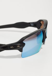 Gafas de sol deportivas negras con un diseño envolvente, que cuentan con lentes en degradado azul y marrón y un acento metálico en la montura.