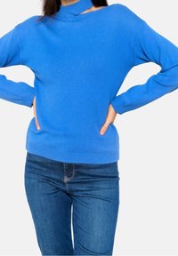 Pull en laine bleue avec un col montant et des manches longues, présentant une taille cintrée et une texture lisse. Porté avec un jean en denim bleu.