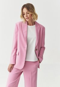 Blazer fucsia su misura con rever a punta, chiusura con un bottone e tasche laterali. Indossato sopra una maglietta bianca, abbinato a pantaloni a gamba larga.