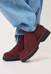 Botas de ante burdeos hasta el tobillo con una suela negra gruesa, cremallera lateral y acabado texturizado, combinadas con unos vaqueros de mezclilla azul claro.