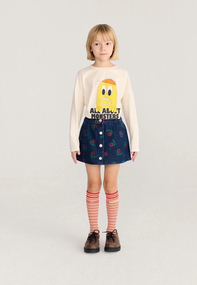 Enfant aux cheveux blonds courts portant un pull crème avec un imprimé monstre jaune, une jupe bleue à motifs floraux rouges, des chaussettes rouges rayées aux genoux et des chaussures marron.