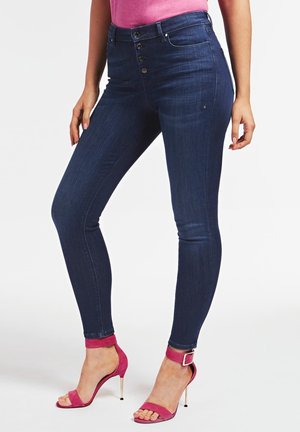 Woman wearing dark blue high-waist skinny jeans and pink high-heeled sandals with ankle straps against white background.