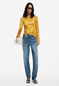 Langärmliges, geripptes Oberteil in Gelb mit Blumenmuster, kombiniert mit hellblauen Jeans aus Denim, dunkelblauben Sandalen und einer kleinen weißen Handtasche.