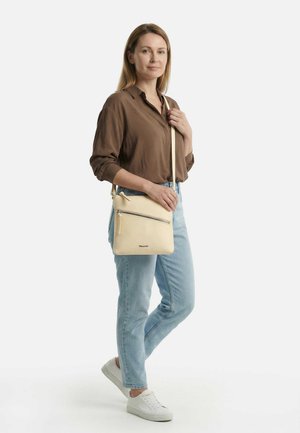 Vrouw in een bruin shirt en lichtblauwe spijkerbroek, met een beige crossbodytas en witte sneakers, staand op een witte achtergrond.
