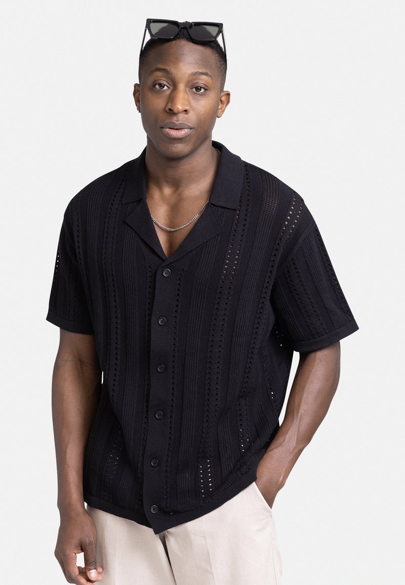 Camisa negra de manga corta con un patrón de punto estructurado, presenta cierre de botones, cuello abierto y acentos texturizados.