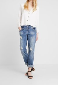 Vit skjorta med knappar, blå slitna denimjeans, uppvikta byxben och svarta sandaler. Skjortan har en strukturerad yta och en något lös passform.