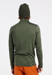 Olivgrünes Langarmhemd mit einem hohen Kragen, aus glattem Stoff gefertigt. Getragen mit einer strukturierten dunkelgrünen Strickmütze.