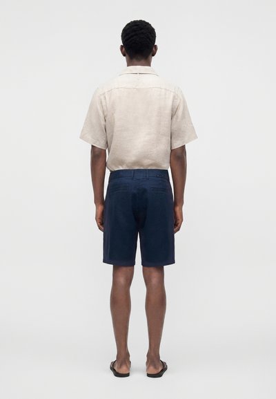 Homme debout de dos, portant une chemise beige à manches courtes, un short bleu marine et des sandales noires sur un fond clair uni.