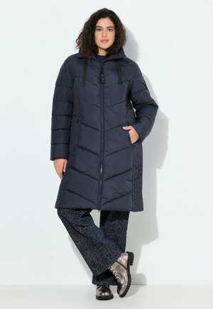 Frau in einer marineblauen gesteppte langen Jacke, dunklen gemusterten weit geschnittenen Hosen und metallischen Schnürstiefeln, vor einem schlichten hellen Hintergrund stehend.