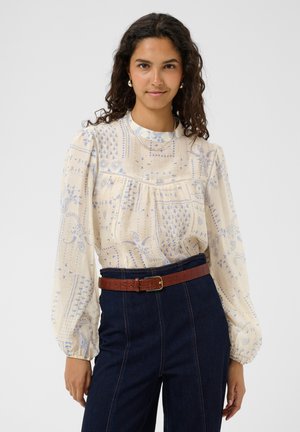 Femme aux longs cheveux bouclés porte un chemisier crème à motifs bleus, un jean foncé, une ceinture marron, des boucles d'oreilles en perles et un fin collier, debout et souriante.