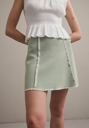 Personne portant un haut blanc sans manches à basque et une mini-jupe texturée vert clair avec une bordure en dentelle blanche, debout sur un fond uni.