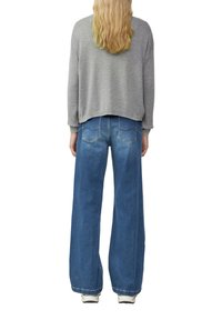 Grauer, verkürzter Sweatshirt mit langen Ärmeln, kombiniert mit weit geschnittenen blauen Jeans. Das Material wirkt weich; die Jeans haben Gesäßtaschen und Nähdetails.