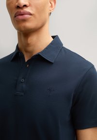 Polo de color azul marino con un collar clásico, tapeta de dos botones y un logotipo bordado en el lado izquierdo del pecho. Textura de tela suave.