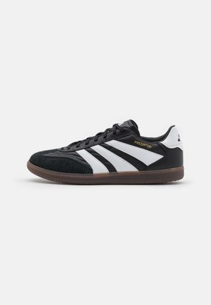 Schwarz-weißer Sneaker mit niedrigem Schnitt und brauner Sohle, drei weißen Streifen an der Seite und "PREDATOR" goldfarbenem Text in der Nähe der Schnürsenkel.